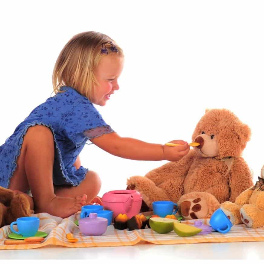 A young child feeds a teddy bear at a picnic.