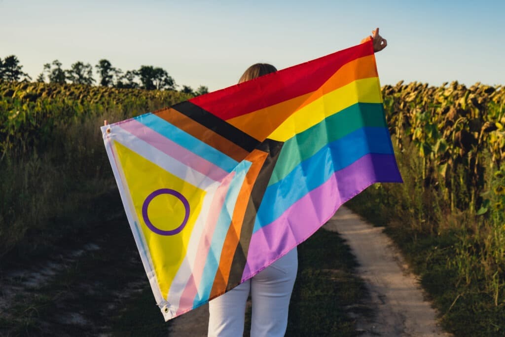 Young woman run in field with Rainbow LGBTQIA flag waving in wind made from silk material on field background.